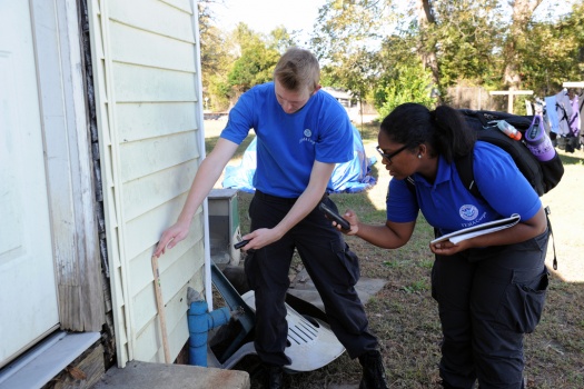Two FEMA Corps members observe the outside of a home. They are holding clip boards to record information and taking a picture of the wall.