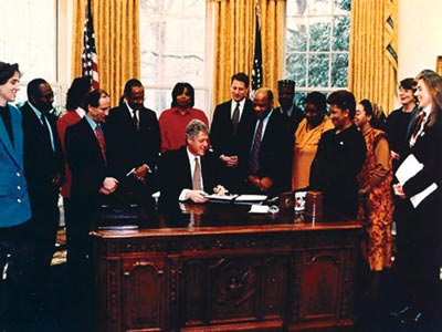 Photo. President Clinton is sitting at his desk in the Oval Office signing a document, with 14 people standing around his desk.