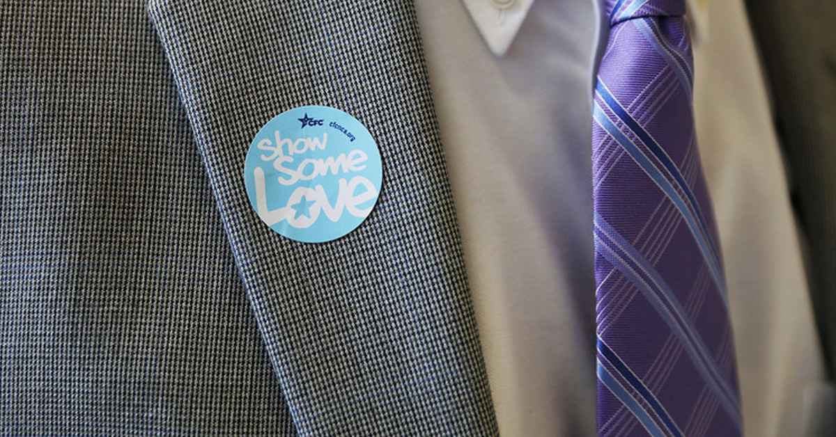 Picture of a gray suit, with a purple tie. On the suit is a blue circle sticker. It says 'Some Some Love'in white  and has the CFC logo at the top in dark blue.