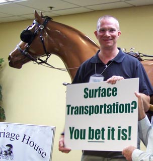 Christopher standing next to a horse while holding a sign that says: Surface transportation: You bet it is!