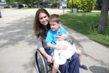 A mother and son, sit outside in a driveway facing forward. Both are smiling. The mother is a wheelchair user. Her son, wearing shorts and sandals, sits on her lap holding a bunny rabbit.