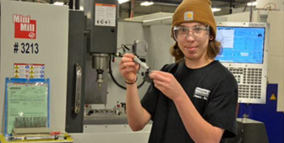 Young man in brown skullcap holding a piece of equipment and standing in front of machinery.