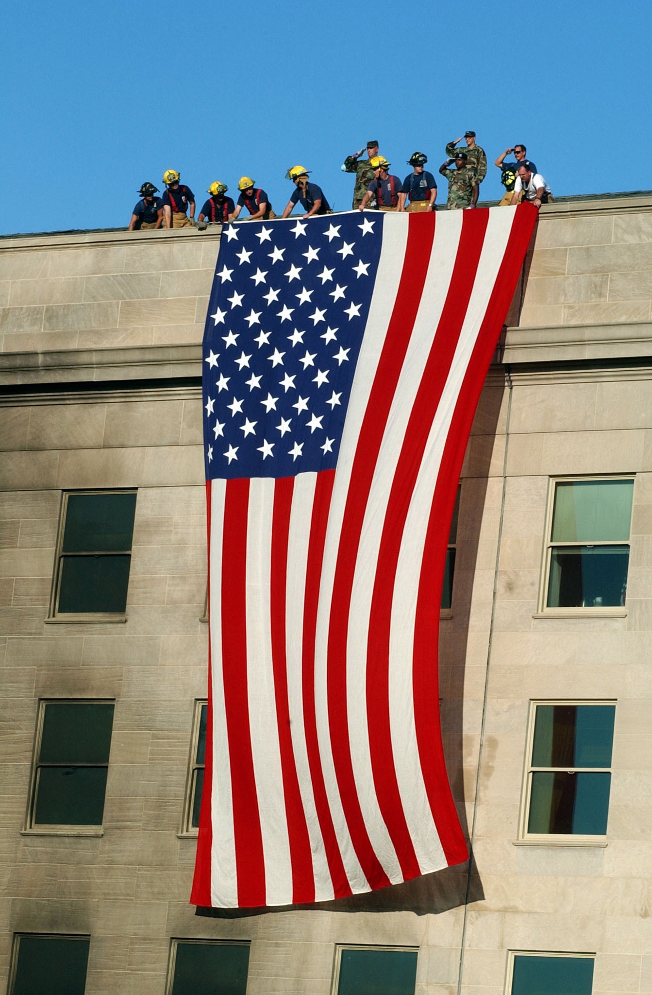 Military service members salute as fire and rescue workers unfurl a huge American flag over the side of the Pentagon, Sept. 12, 2001, during rescue and recovery efforts following the 9/11 terrorist attack. At about 9:40 a.m., a hijacked commercial airliner, originating from Washington’s Dulles airport, was flown into the south side of the building. Navy photo by Petty Officer 1st Class Michael W. Pendergrass