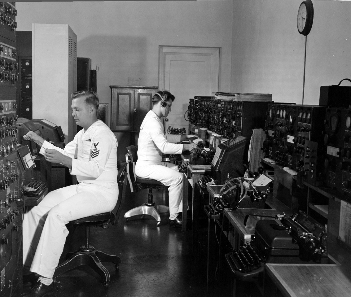 At left RM1C William G. Turpin reads radio-teletype tape just received at Naval Radio Station, Cheltenham, Maryland, from the Commander Operational Development Force, ready for automatic retransmission to Radio Washington. At right, RM3C D.F. Wellner handles manual ship-shore radio circuit. U.S. Navy photo.