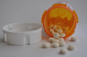 An orange prescription bottle lies on its side with its white cap next to it. Small pills spill out from the bottle.