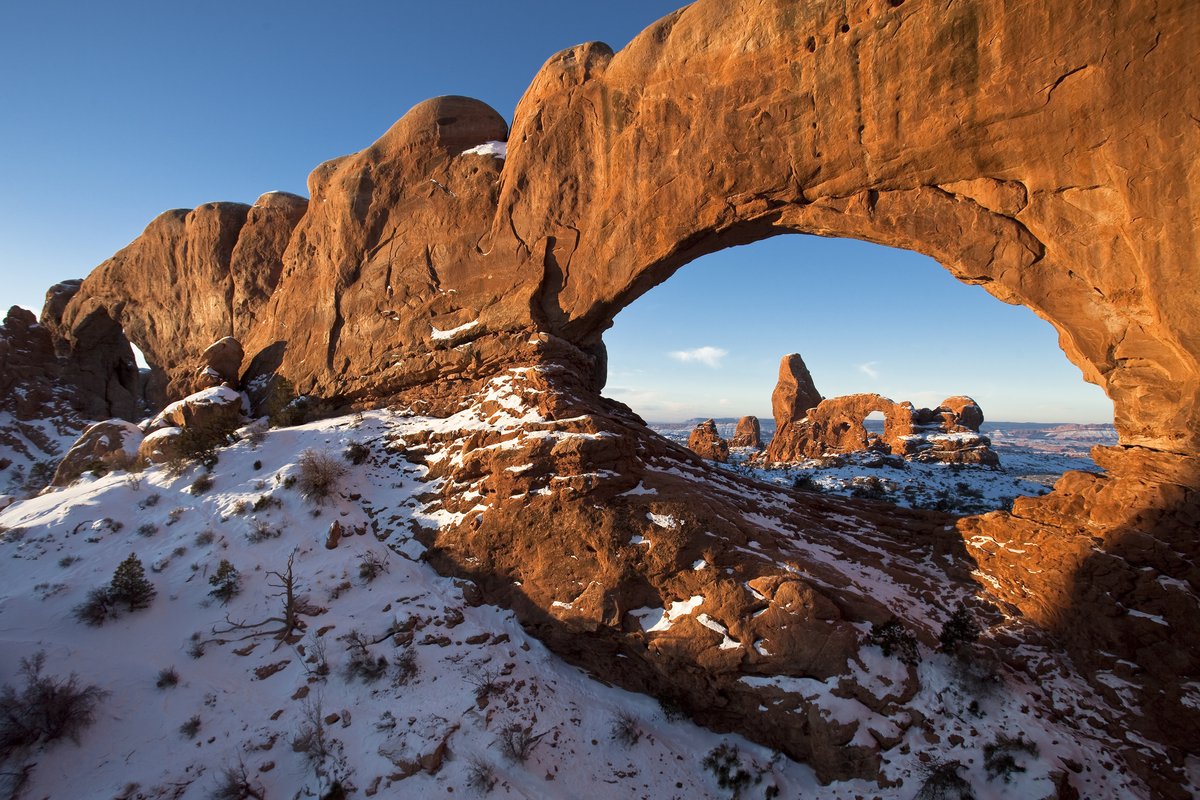A rock formation in the distance is seen through a hole in a closer rock wall. The landscape is dusted with snow and lit with morning light.