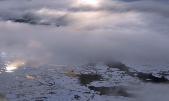 Clouds drift in beams of sunlight above large icebergs floating on the ocean.