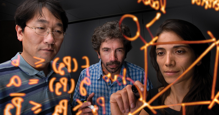 A photo of a woman and two men writing calculations on a clear screen.