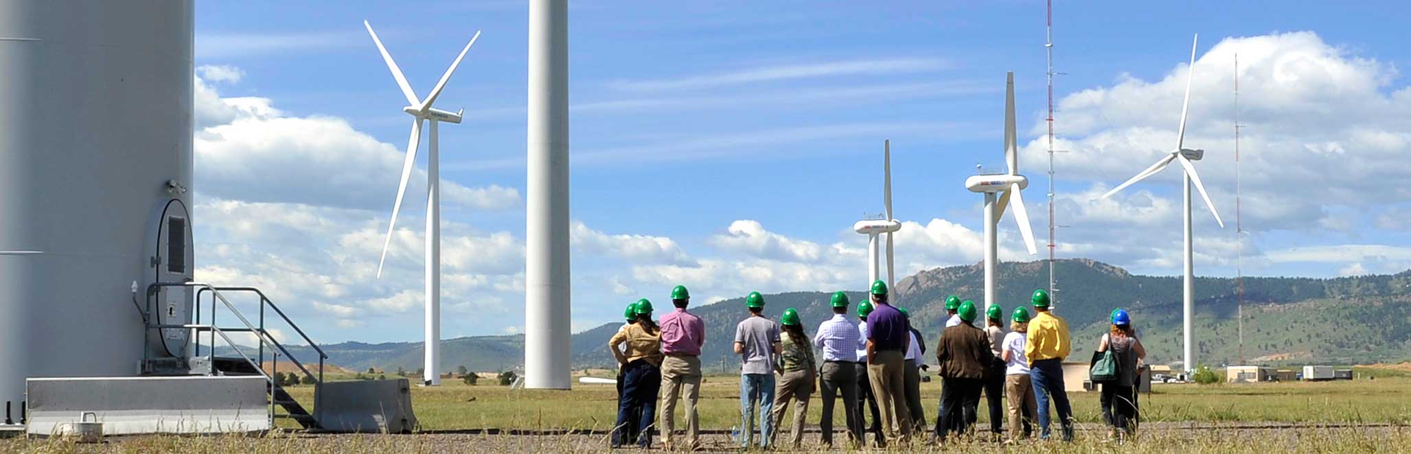 A group of NREL Energy Execs gather around the bottom of a wind turbine with blue skies and fields in the foreground.