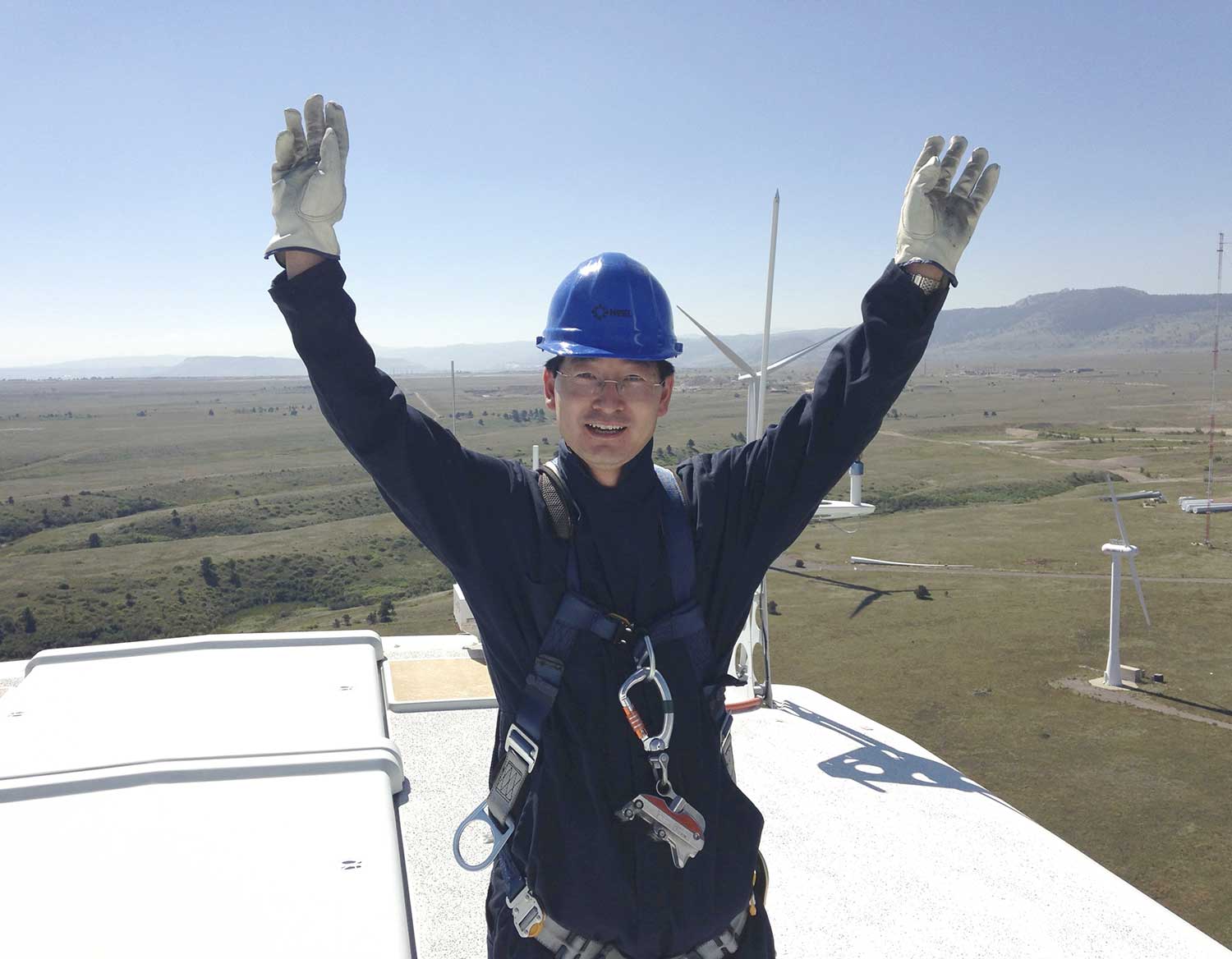 A photo of a man wearing a hard hat and a harness standing on top of a wind turbine throwing his hands in the air.