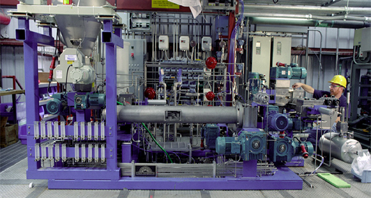 Photo of a 200 kg/day Small Horizontal Reactor in an industrial plant setting. It is the smallest of four continuous chemical pretreatment reactors and is used for piloting to the larger throughput systems. A man in a hardhat and safety glasses stands to the right the reactor.