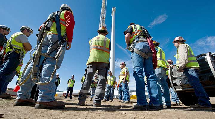 A photo of several people in safety vests and hardhats standing near a land-based wind turbine shaft with its blades on the ground in the background.