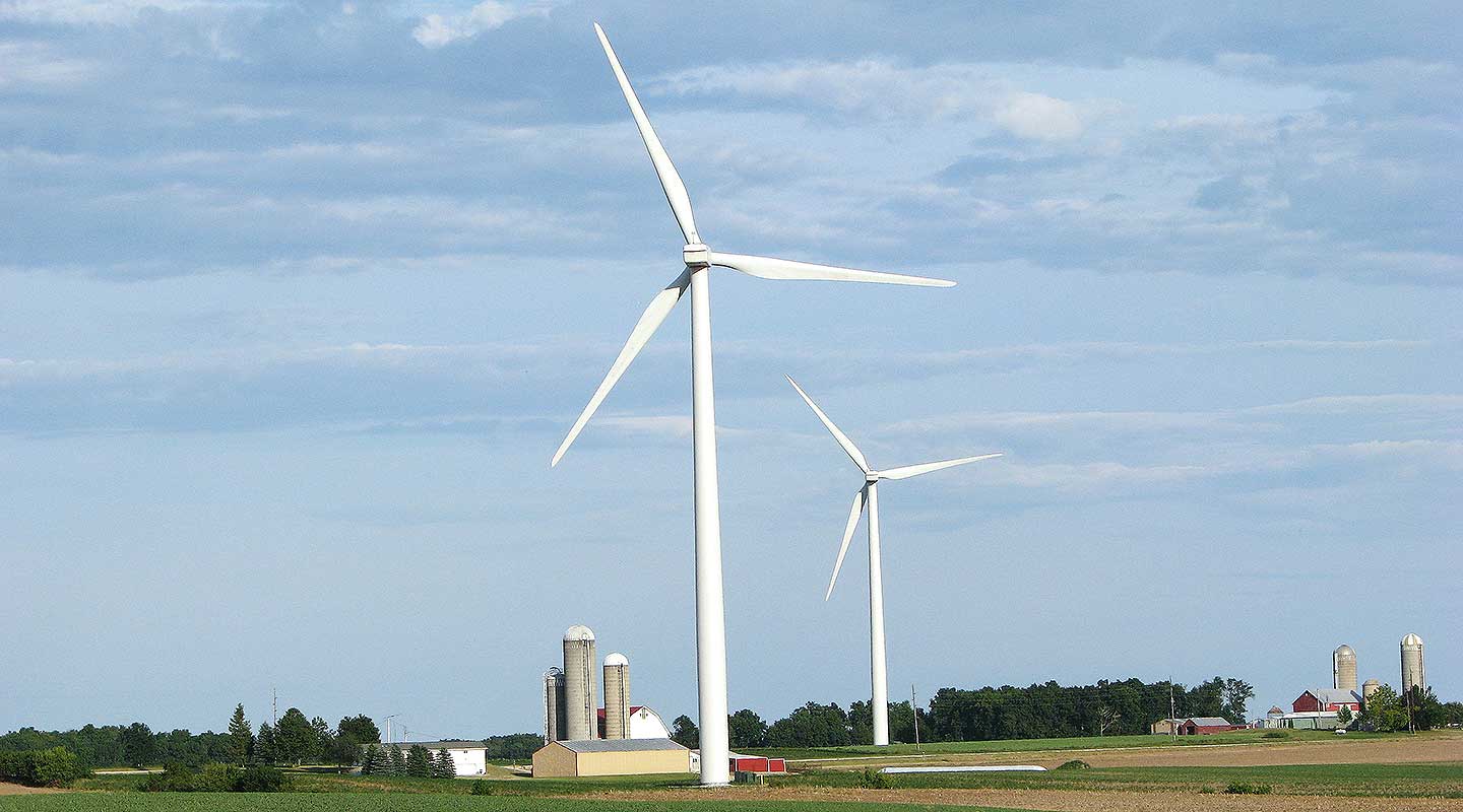 A photo of two large wind turbines in a green field with a farm and silo next to the turbines.