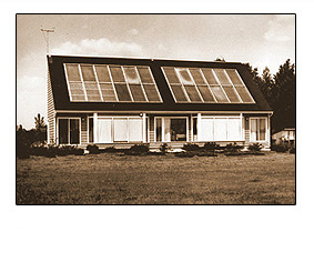 Black and white photo of  a  slanted roof on a white house. A TV antenna is displayed on the left-hand side of the rooftop, along with 12 rows of solar panels , and a row of bushes in front of the house.