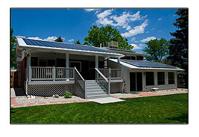 Photo of a white house with a slanted roof, and several vertical , rectangular-shaped panels displayed on top of the roof. Blue sky, and bright-green trees fill the background of the photo.