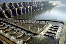 Water flows down a stairstep aqueduct from the top of a large river dam to the bottom. The aqueduct makes two 90-degree turns to the left on the way down before reaching the river at the base of the dam.