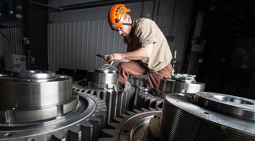 Photo of a man wearing an orange helmet and measuring a pin on one of three large gears, which are part of a drive train.