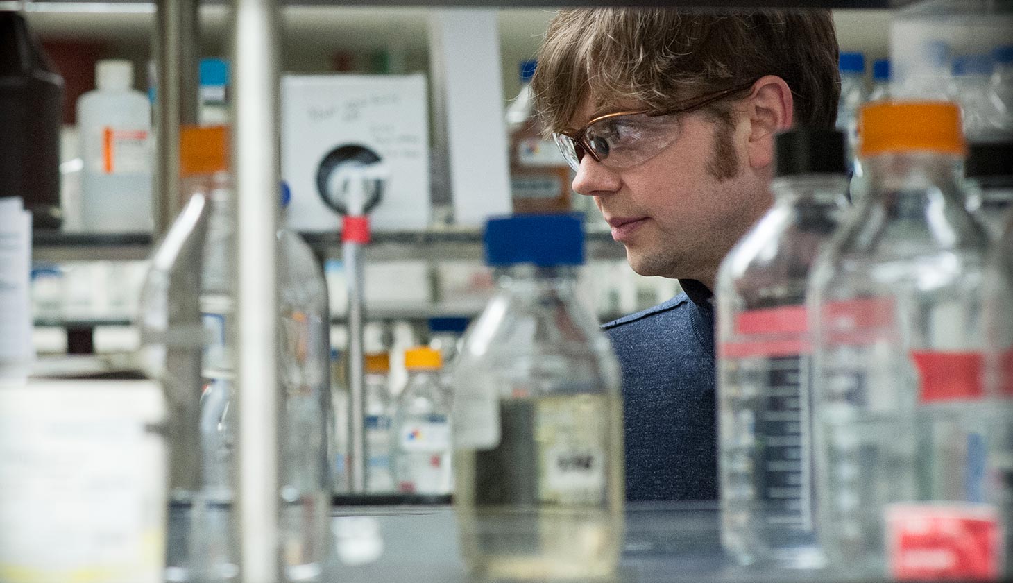 Man, Gregg Beckham, in lab behind shelf of beakers and jars