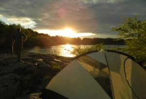 A camper stands on the shores of a campsite with late afternoon sun shining over the river. A tent has been pitched in the foreground. 