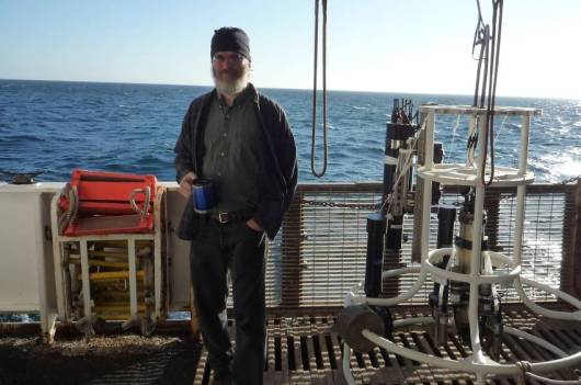 Dr. Steve Pierce  at his work station and standing next to the CTD on a bright sunny day in the Northern Pacific Ocean. 