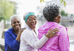 A group of three happy, senior African American women standing together at the park. Two of them are looking at the camera and laughing. The main focus is on the woman in the middle.