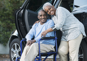 A happy senior African American couple outside a car. The man is sitting in a wheelchair and his wife is standing beside him with her arm around his shoulder.