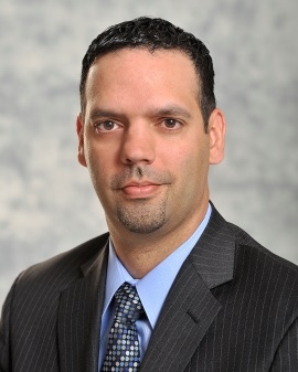 Photo of man with short-cropped black hair wearing gray pin-striped suite, blue shirt and tie.