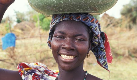 A smiling woman in West Africa carries a large bowl on her head.