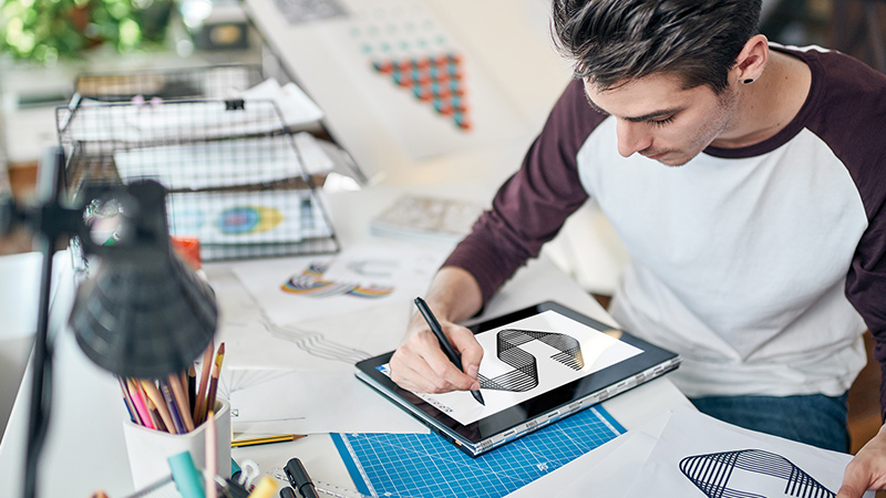 Man drawing geometrical letter S on on a 2-in-1 while sitting at desk surrounded by graphic design materials