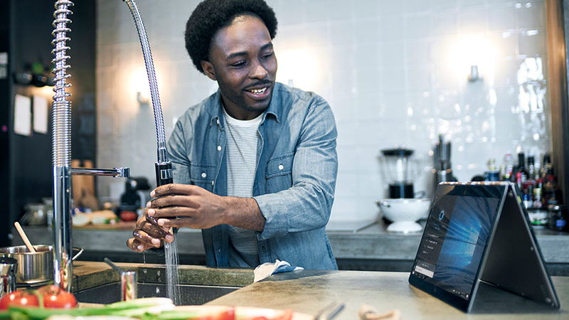 Man looking at Cortana on a 2-in-1 device with Cortana on screen while running water in the kitchen sink