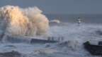 Huge waves crash against the coast. A lighthouse can be seen