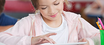 Little schoolgirl smiling while using digital tablet at desk in classroom