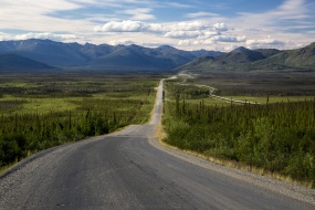 A motorcylcist and truckers pass each other on the rough, dusty, unpaved Dalton Highway approaching the Brooks Range. The Trans-Alaska Pipeline parallels the highway.