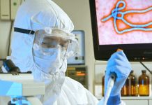 عالم يرتدي سترة وقاية كاملة داخل مختبر وخلفه شاشة تظهر عليها صورة لفيروس إيبولا. (© AP Images)