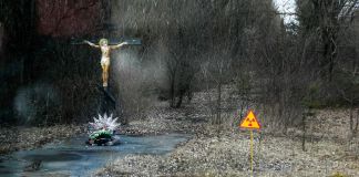 Une statue du Christ en croix et un panneau Danger radiations au bord d'une route à l'abandon, recouverte de feuilles et entourée d'arbres. (© AP Images)