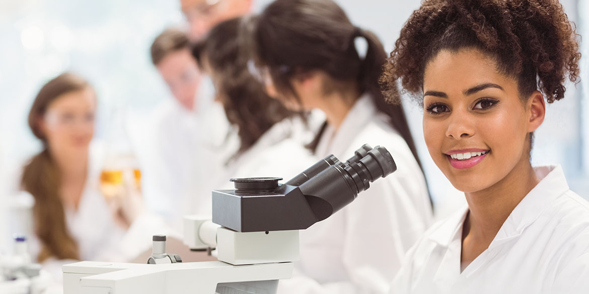 A woman in a lab coat smiles next to a laboratory microscope.