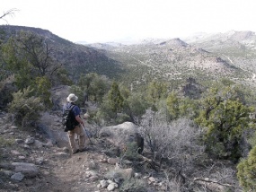 A hiker pauses on a trail that runs on ahead of him into some mountains in the distance.