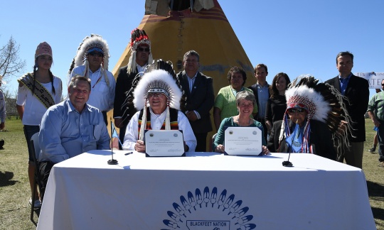 Secretary Jewell and three other people sit at a table holding up a signed document. Other people pose in a line behind them, some wearing tradition Indian headdresses. The event is outside on a bright day.