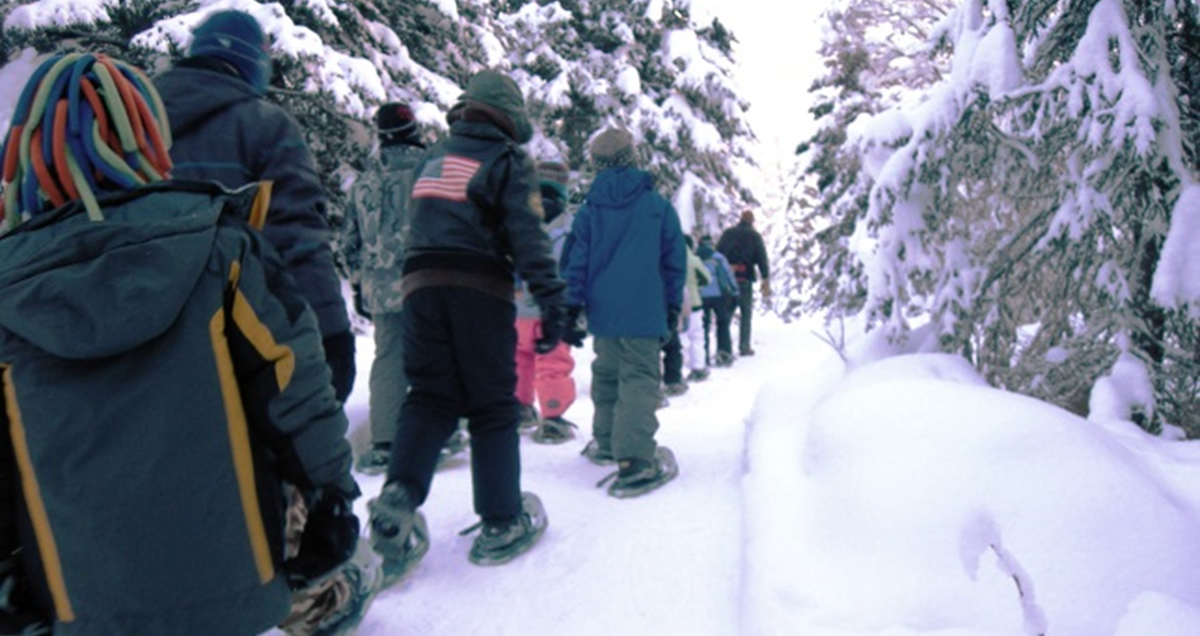 A line of students on snowshoes follows an instructor through the snowy forest on Campbell Tract.