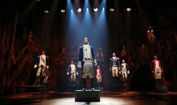 A man dressed in an American eighteenth century costume stands under the spotlight on a large stage.