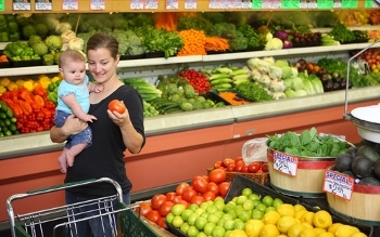 Young mother with baby selects items in produce aisle of grocery store