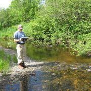 USGS technician monitoring a low flow in a stream while standing on a pebble island in stream in summer.