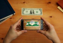 Hands holding a phone displaying the White House and dollar bill on the table (White House)