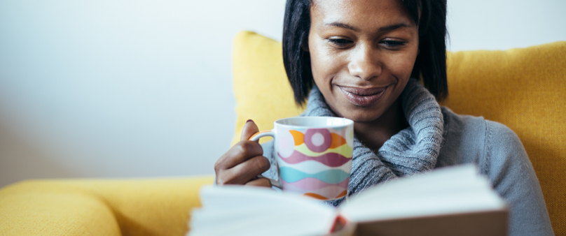 Photo of a woman reading a book with a mug of tea. She's sitting in a yellow chair.