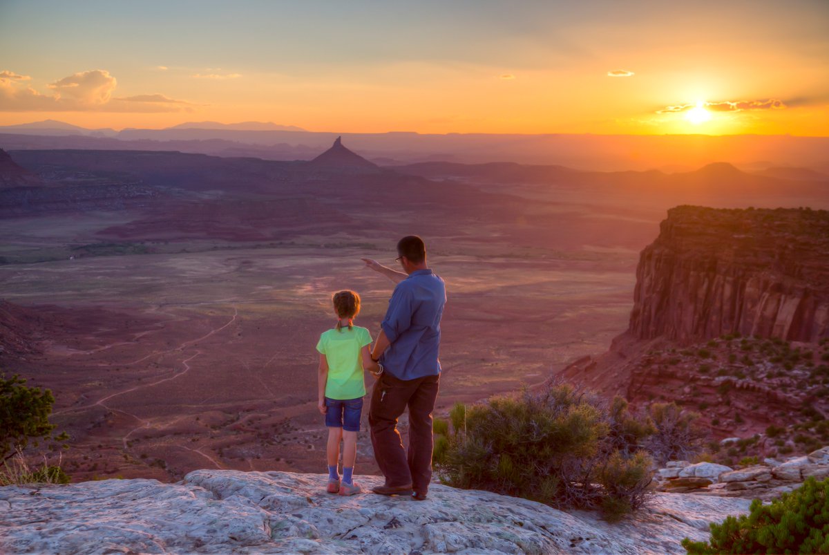 A man and a girl look out across a valley at sunset as he gestures toward a peak in the distance.