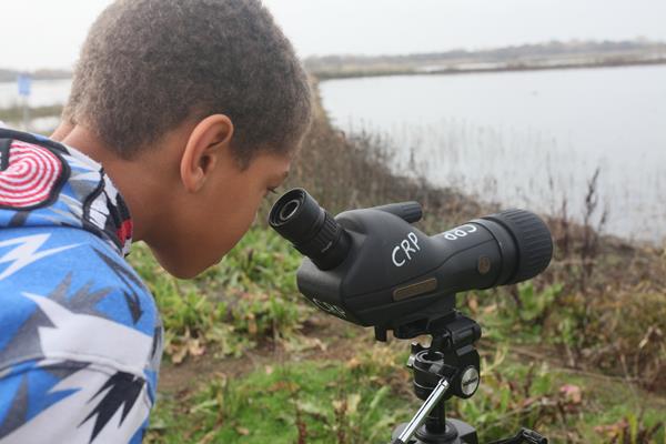 A boy looks through a telescope across a lake on a cloudy day.