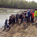 big group of kids standing on a river bank watching a science demonstration