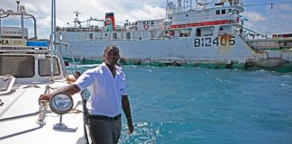 Homem se segura em parapeito de barco em alto mar; embarcação maior ao fundo (© kashfihalford.com)