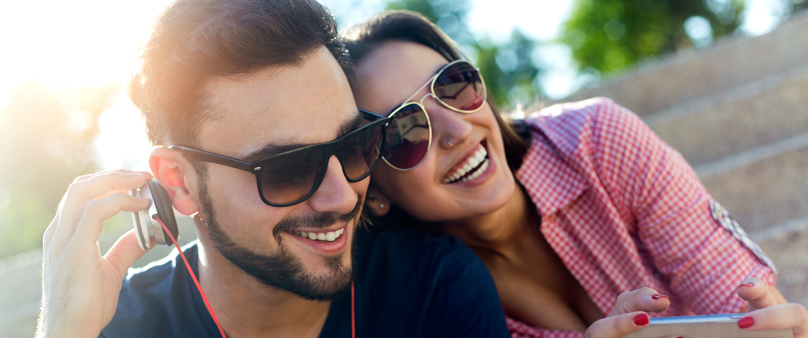 Photo of a man sitting outside on steps listening to headphones. His girlfriend sits next to him, holding her phone. 