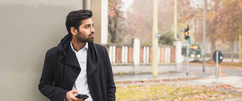 Photo of a man leaning against a wall in a neighborhood. He's holding his phone.